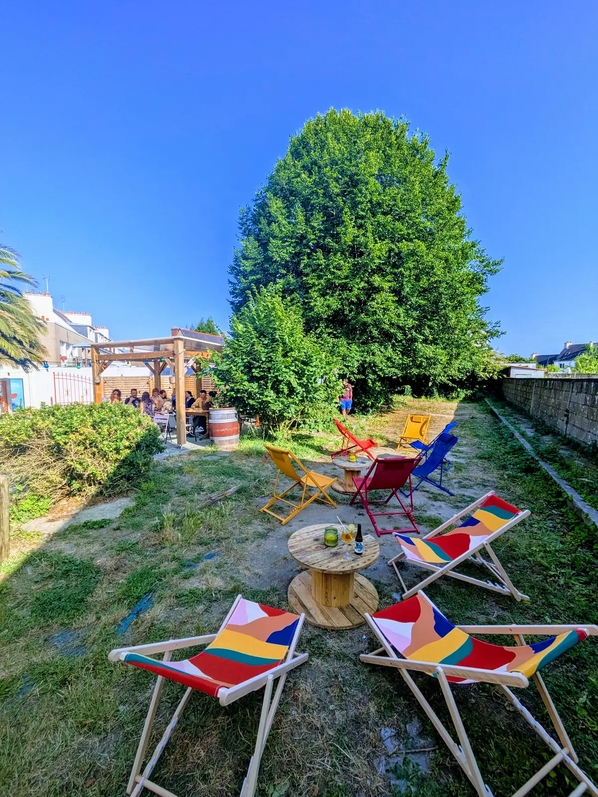 Les transats colorés du jardin d’À Ton Aise à Lorient, autour de tables en touret, sous le grand tilleul